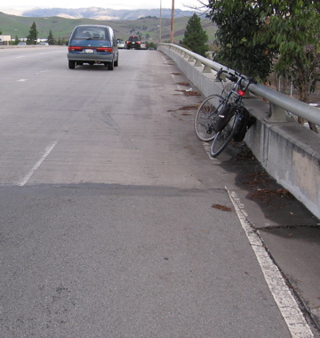 But here (top of bridge), the 8-foot shoulder was eliminated in 2003. The former shoulder line is still visible. Note bicycle. The new shoulder line is TOO NARROW for bicyclists and pedestrians. (looking east, eastbound side)